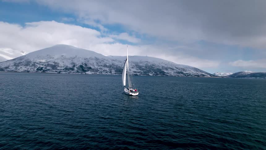 A sailboat sails the ocean with the shore ahead and snowcovered mountains in the distance on a cloudy day