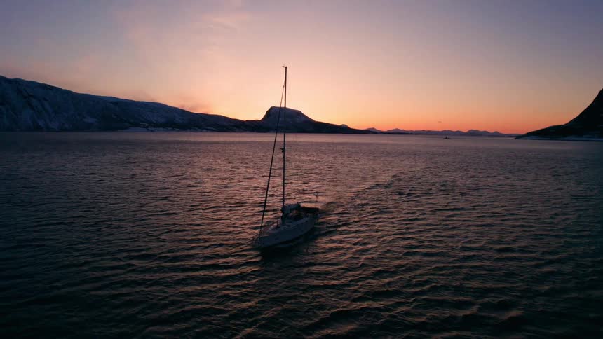 Sailboat travels under power leaving a wake while sunlight shines behind coastal mountains on the horizon