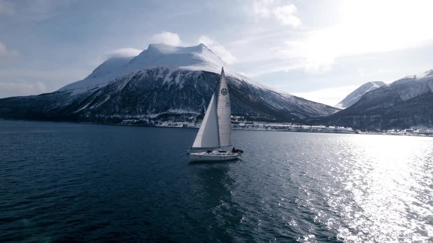 The video features a beautiful white sailboat gliding across a tranquil body of water with the suns reflection shimmering on the surface majestic snowcapped mountains rise dramatically in the background under a partially cloudy sky