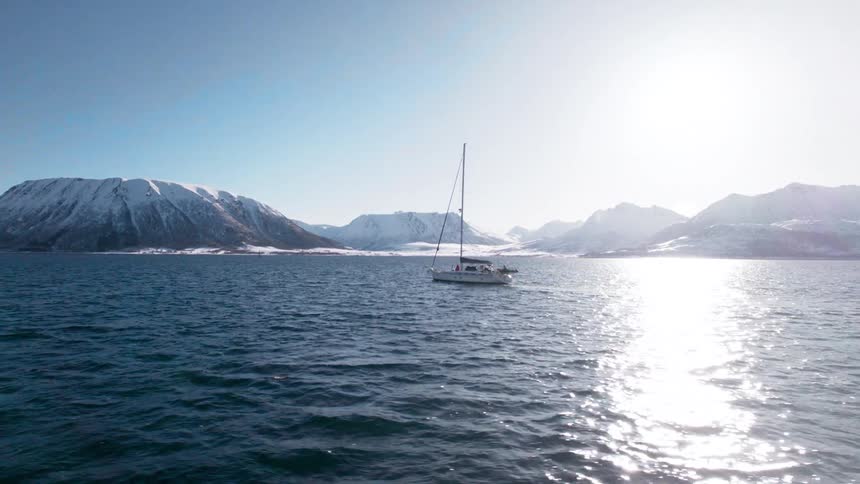 A sailboat sails the ocean at midday with the shore ahead and snowcovered mountains in the distance
