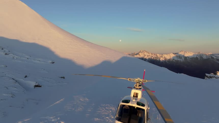 A helicopter rests on a snowcovered mountain ridge under golden sunrise light with distant peaks visible under a clear sky