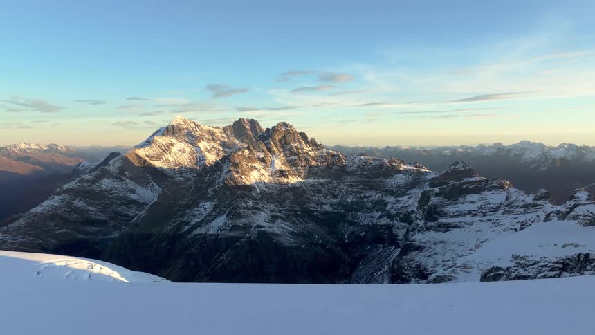 The video showcases a magnificent mountain range under a clear sky with the sun casting a golden glow on the snowcapped peaks in the foreground a pristine expanse of snow extends towards the viewer hinting at a highaltitude vantage point