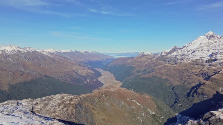 A panoramic view of a wide mountain valley under a vivid blue sky bordered by snowy peaks and forested slopes