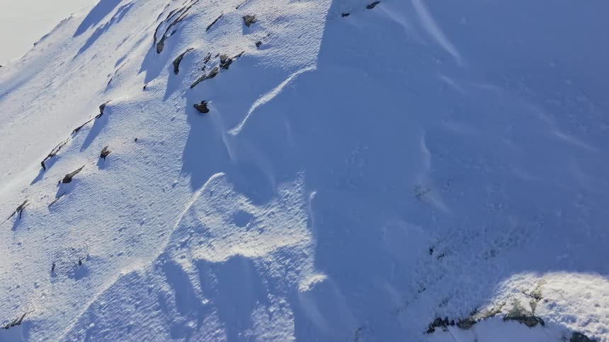 The video captures an extreme highangle shot of a skier on a vast snowcovered mountain with snowladen boulders and jagged rock formations creating a dramatic landscape the scene is dominated by bright whites and deep blues suggesting a clear