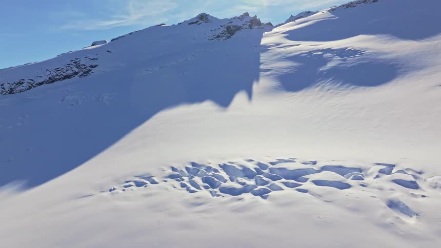 A pristine sunlit snowfield stretches toward rugged peaks with sharp mountain shadows casting deep blue shapes across the untouched surface