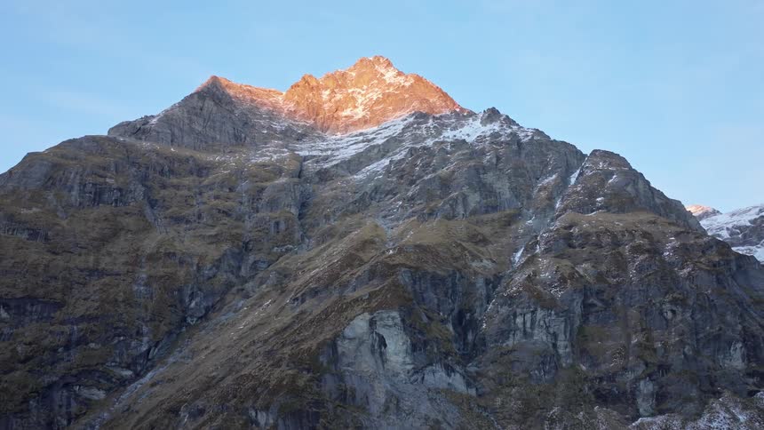 A rugged mountain face is streaked with snow and rock crowned by a sunlit summit glowing warmly against the pale blue sky