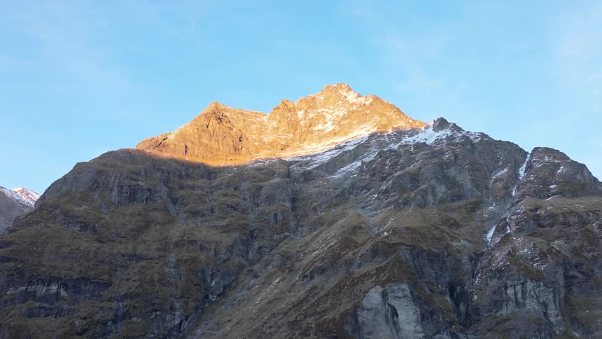 The video features a majestic mountain peak with its upper slopes bathed in golden sunlight and its lower half covered in a mix of rugged rock and sparse vegetation the sky above is clear and blue completing a dramatic natural landscape