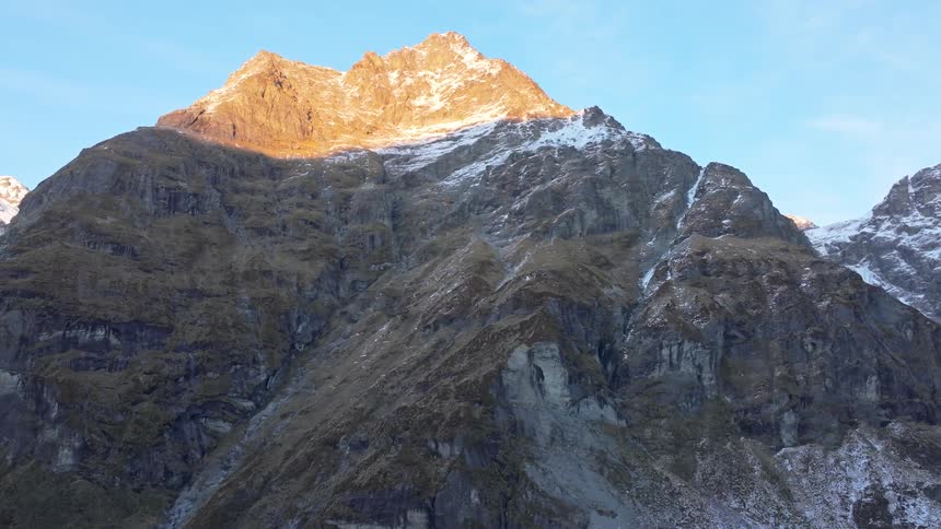 A towering mountain peak dusted with snow glows in the golden sunlight against a clear blue sky