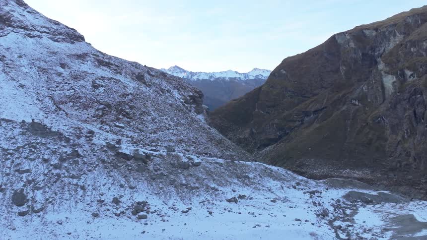 The video shows an expansive mountain valley with scattered snow on the slopes and a stream winding through the foreground towering rocky mountains rise on either side of the valley with more snowcapped peaks visible in the distance under a clear sky