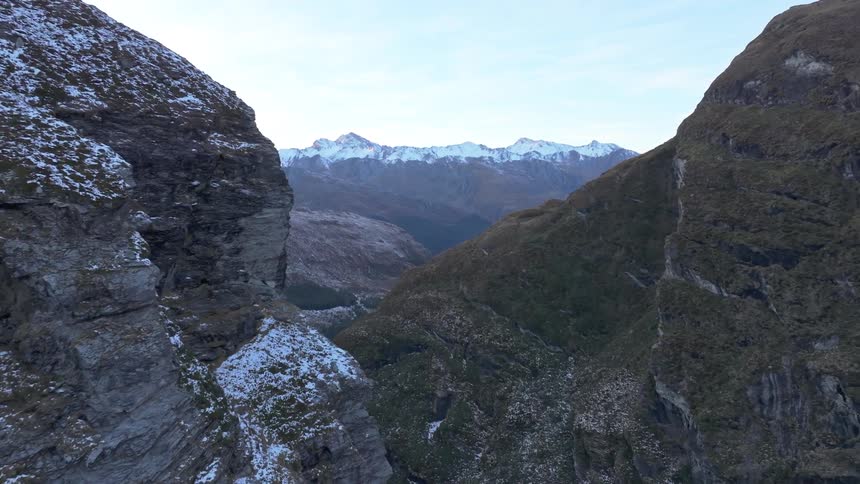 The video shows an expansive view from a mountain pass revealing two steep rocky slopes framing a deep valley in the distance a majestic range of snowcapped mountains stands against a light sky completing the vast rugged landscape