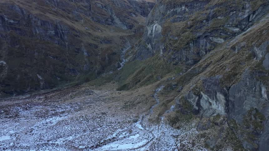 A narrow rocky valley shows traces of snow and a winding stream bed flanked by steep mossy cliffs under fading evening light
