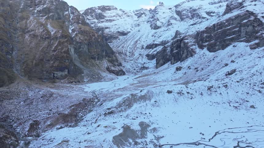 A wide rugged alpine basin lies between towering snowcovered cliffs with icy streambeds winding through the partially frozen terrain