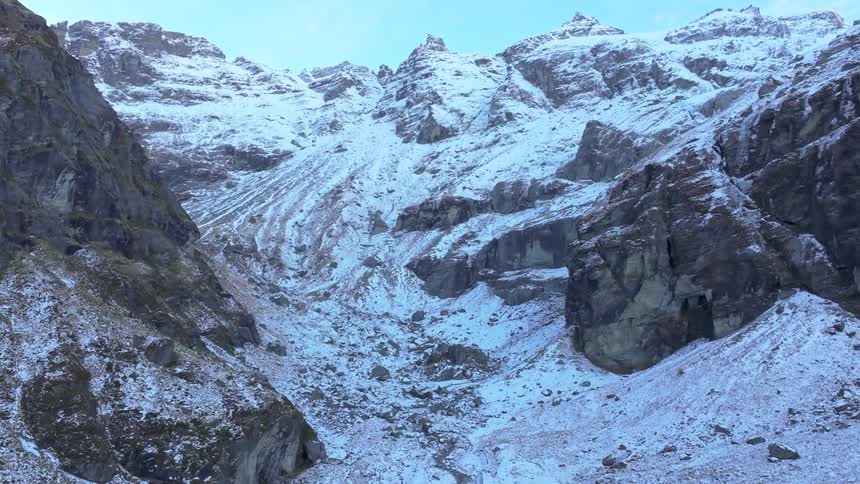 The video presents a striking view of a narrow snowcovered valley or gully framed by towering rugged mountains the peaks and upper slopes are heavily dusted with snow contrasting with the dark exposed rock faces lower down all under a clear
