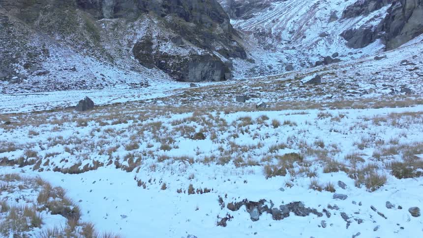 A highaltitude valley shows sparse snow cover over golden grass and scattered rocks framed by steep rugged mountain slopes
