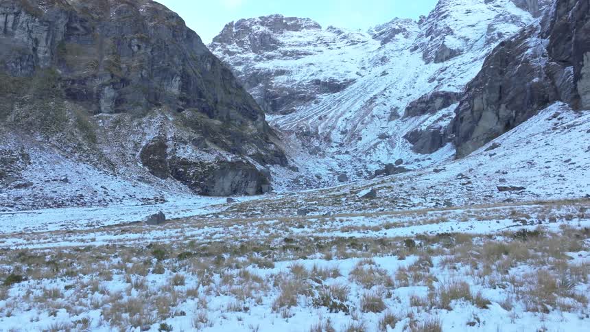 The video shows a wide shot of a valley covered in a light layer of snow with patches of dry yellowbrown grass poking through towering rocky mountains with snowdusted peaks flank both sides of the valley meeting at a distant snowcovered mountain
