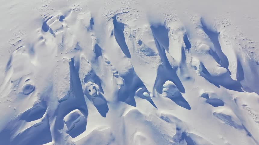 This video provides a mesmerizing aerial view of snowcovered terrain showcasing intricate patterns of shadows and light created by the undulating snowdrifts and ridges the stark white and cool blue tones highlight the rugged untouched beauty of the sn