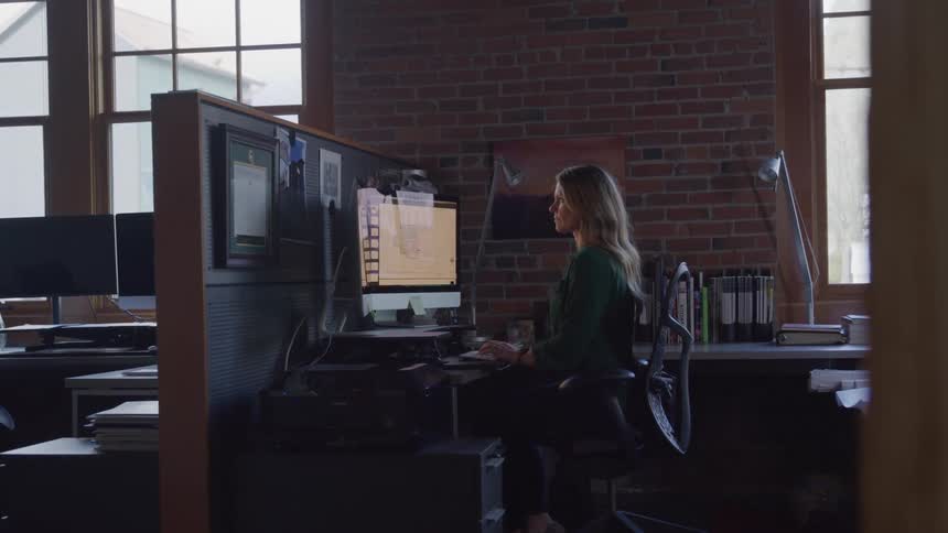 A woman sits at a desk in an office working on a computer with multiple monitors with brick walls and large windows in the background