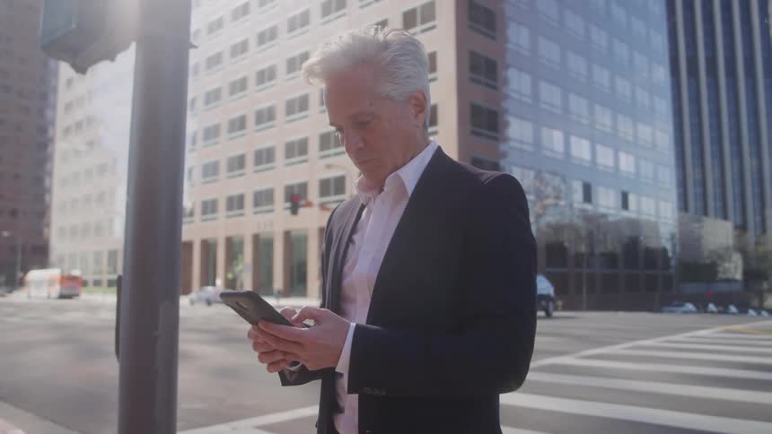A whitehaired man stands on a city sidewalk while looking at his phone