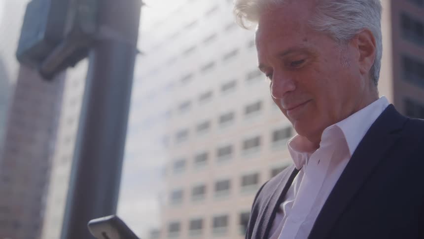 A man wearing a suit scrolls through his phone with a smile on his face as he walks down the street at midday