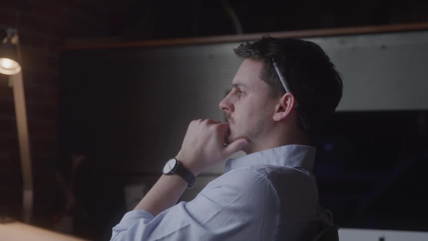 A young adult man likely an architect or designer with a pencil behind his ear and a watch on his wrist looks thoughtfully to the left resting his chin on his hand while a desk lamp illuminates a desk in a dimly lit office