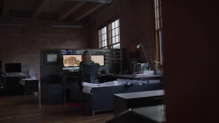 The video opens from behind a dark foreground element revealing a spacious office with exposed brick walls and large windows where a blondehaired woman is seated at a desk looking at two computer monitors displaying architectural designs