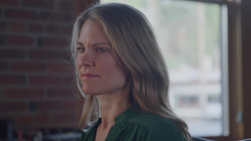 A blonde woman with wavy hair and a green blouse looks thoughtfully to the side sitting near a window with natural light