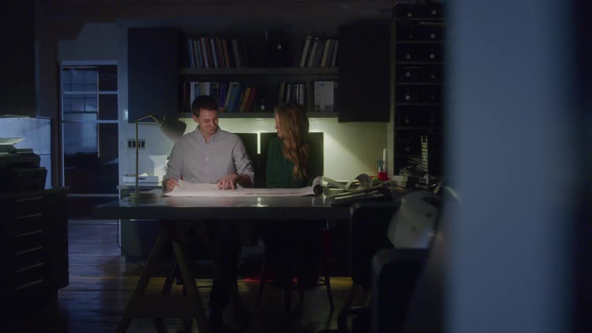 Two coworkers collaborate late into the evening reviewing documents in a warmly lit booklined office