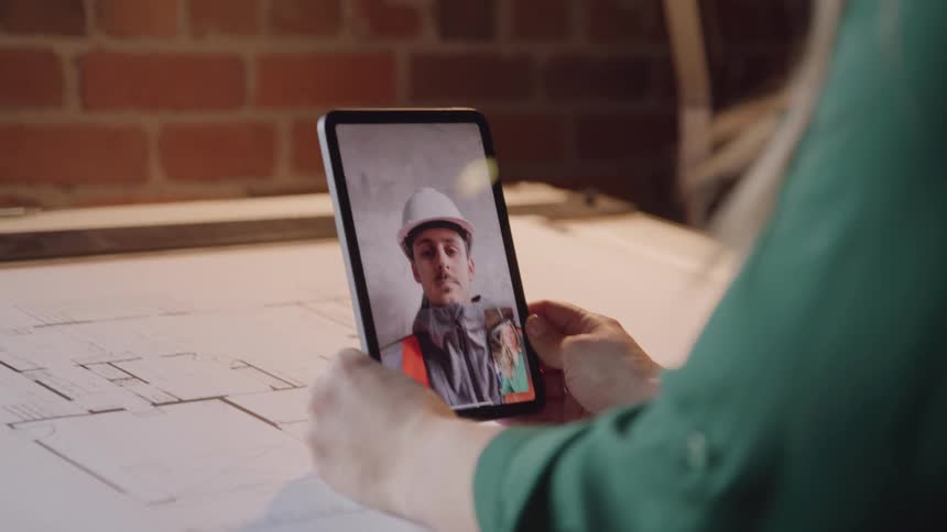 An architect holds a tablet over an architectural drawing on the table in front of her as she engages in a video call with a construction worker wearing a hard hat and red vest