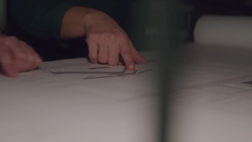 This video provides a closeup view of hands pointing to and interacting with a large architectural or engineering blueprint unfurled on a table the focus is on the details of the plans and the collaborative or instructive nature of the gestures