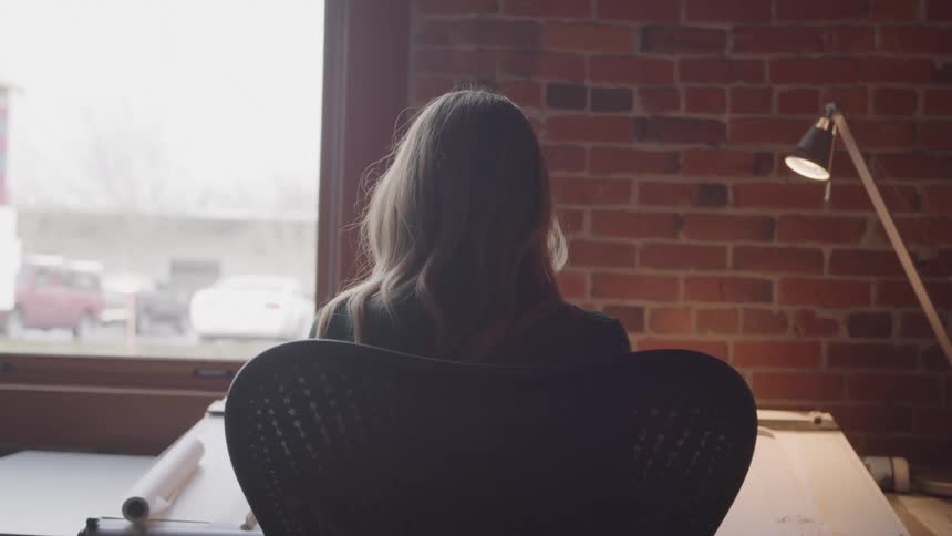 A woman sits alone in an office chair facing a large window backlit by daylight with brick walls and a desk lamp nearby