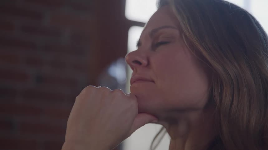 This video shows a side profile of a woman with light brown hair appearing thoughtful or pensive with her chin resting on her hand the soft lighting highlights her contemplative expression against a blurred background that includes what looks like a wi