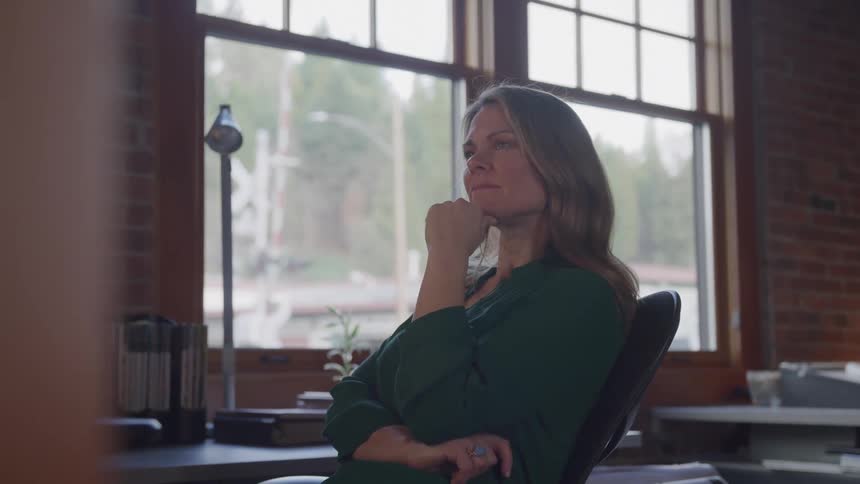 A young adult woman with long hair wearing a dark green shirt sits at a desk and looks thoughtfully out a large window that reveals trees and other buildings