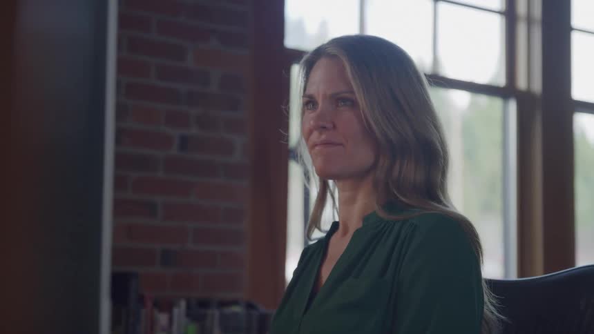 The video opens with a view of a dimly lit room with a window revealing a blurred outdoor scene a woman with long light brown hair then comes into focus appearing thoughtful or contemplative as she looks offcamera to her left