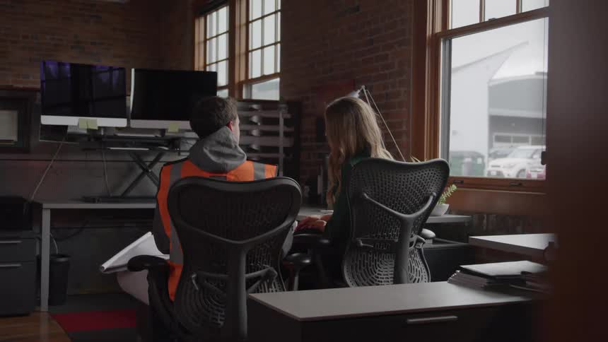 A man in an orange safety vest sits next to a female talking at a workstation with dual monitors partially seen through an open doorway