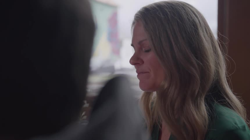 A woman with long blond hair talking to another persons with a thoughtful expression as light comes from a window in the background