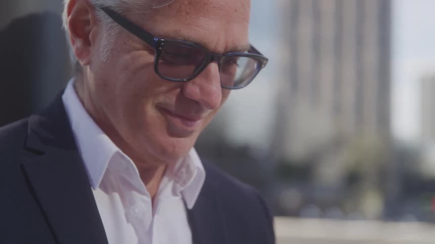 A man in a suit and sunglasses looks down with a subtle smile standing outdoors in an urban setting