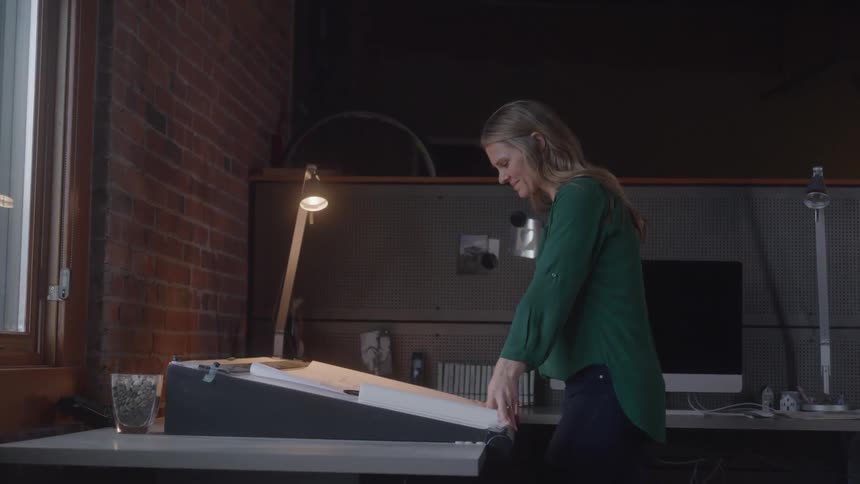 A young woman with long hair wearing a dark green shirt leans over a tilted drafting table with papers on it illuminated by a desk lamp in an office with a brick wall and a window