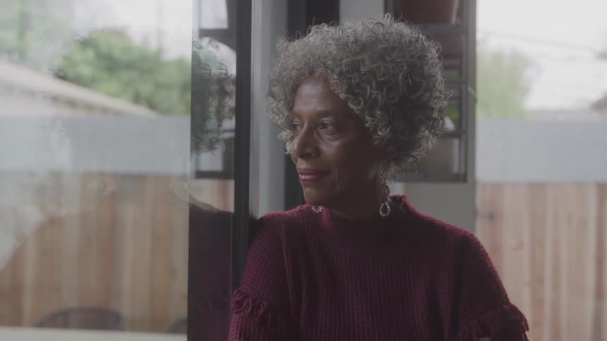 An elderly black woman with gray hair wearing a red dress stands and rests beside a glass window indoors at midday gazing outside