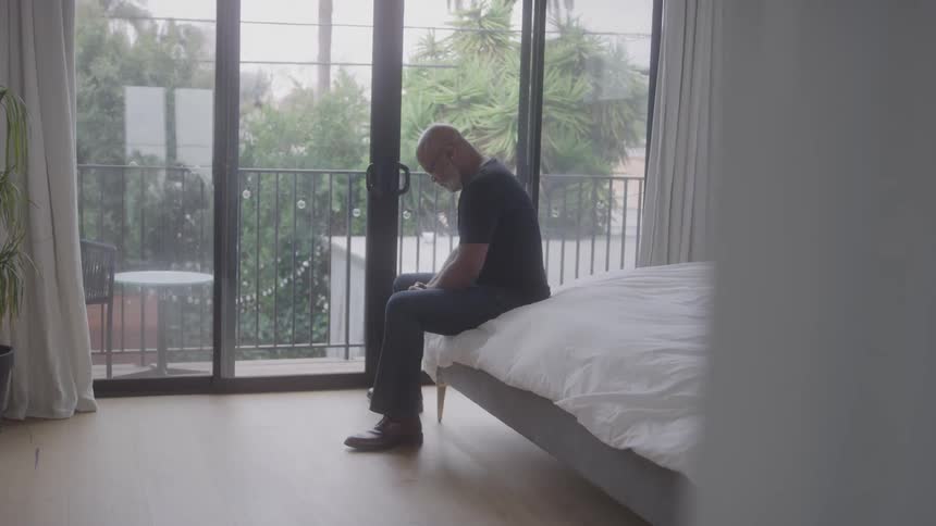 An elderly black man contemplates while sitting at the edge of a bed in a bedroom during daytime