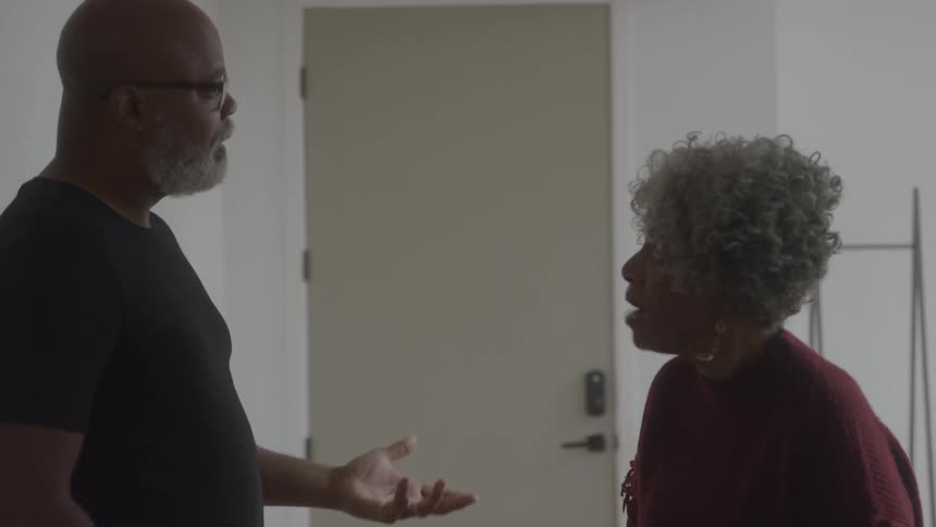 An elderly couple argue intensely in a room near a door