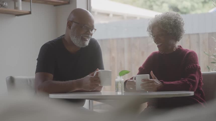 An elderly couple sits at the kitchen table smiling and holding mugs