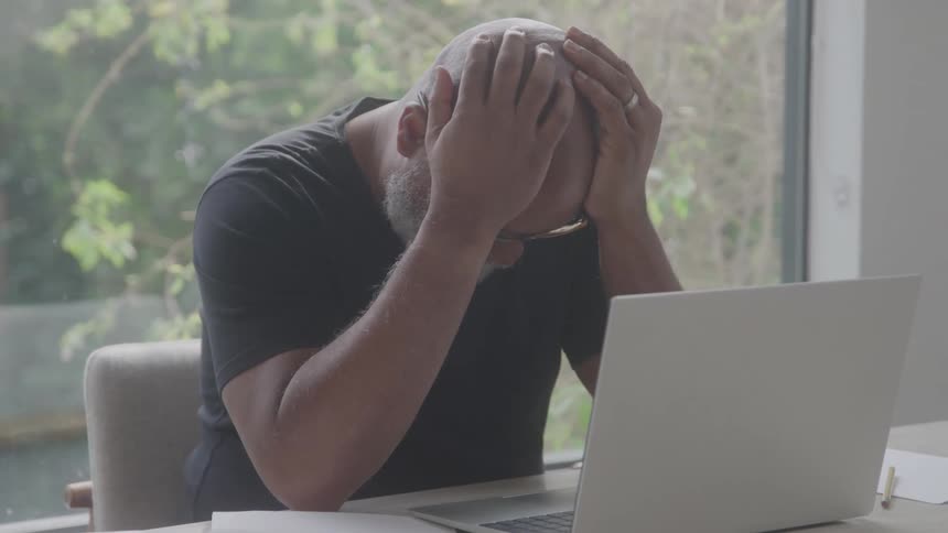 A man sitting at a table with his laptop holds his head in his hands
