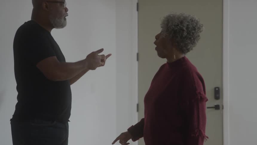 A man and a woman are standing in a room with white walls and are arguing