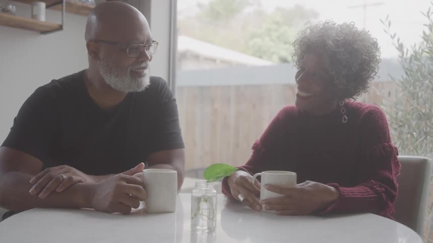 An elderly couple sits at a kitchen table smiling and chatting over mugs of coffee
