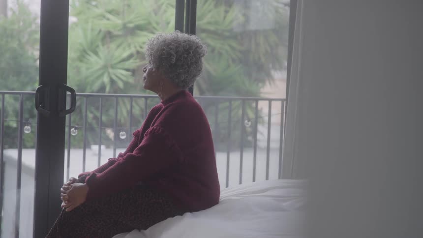 An elderly woman is gazing out the balcony door while sitting on a bed