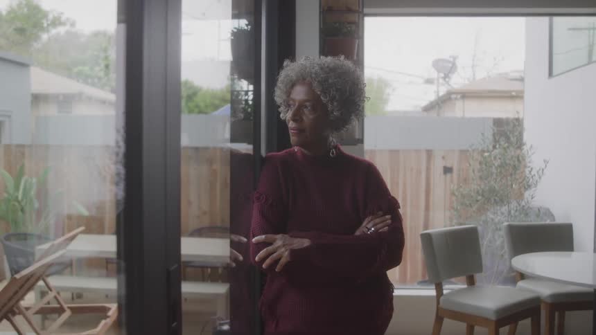 Older woman leans against the glass and looks out into her backyard