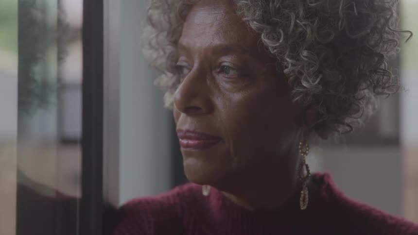 Elderly woman leans near a reflective window and thinks while looking out the window