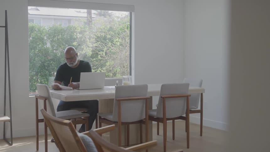 An elderly man sits in the dining area working on a laptop at midday with trees visible outside through the glass window