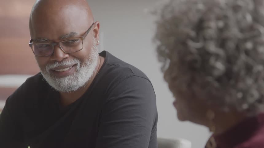 A smiling black man with a white beard and glasses looks warmly at a black woman during their conversation