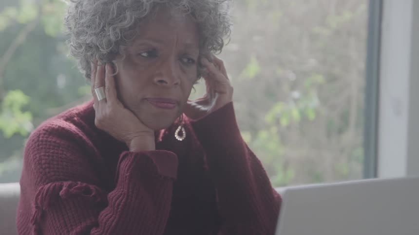 An older adult woman with curly grey hair wearing a red sweater and earrings looks at a laptop with a concerned expression then puts her hands to her head as if in distress